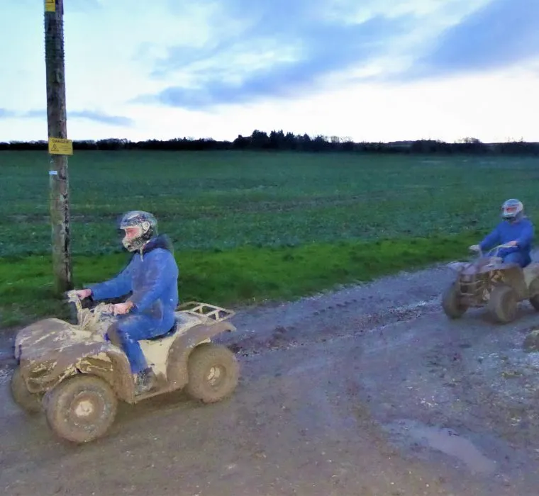 Two quad bikers in blue overalls on muddy track at dusk